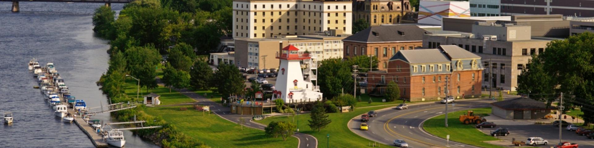 Buildings near a river. There is a jetty with a row of boats on it. There is also a white lighthouse near the coast of the river.