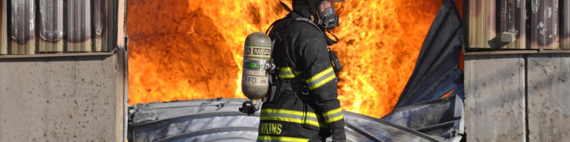 Firefighter stranding in the foreground while there are flames coming out of a structure in the background.